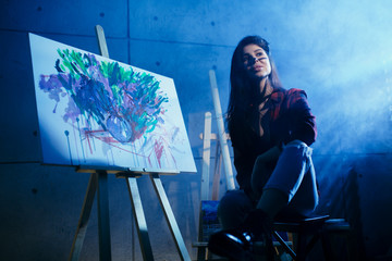 Beautiful brunette girl artist sitting in a dark Studio near the easel with painted watercolor abstract painting.