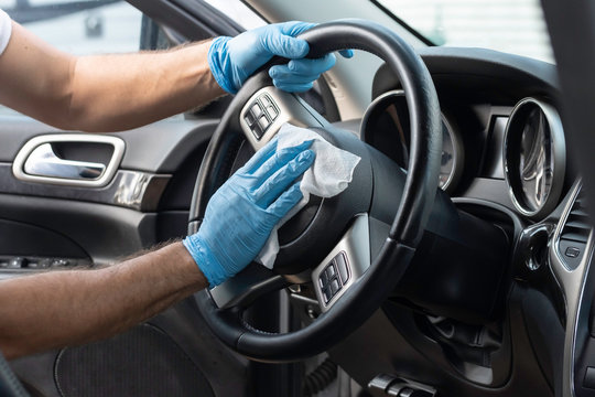 A bearded brown haired man wearing a baseball cap uses alcohol wipes to sterilize the front driver seat, steering wheel, and surrounding areas from any lingering virus strains or germs that are inside