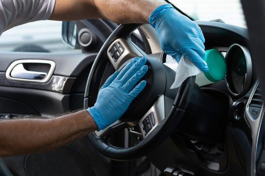 A bearded brown haired man wearing a baseball cap uses alcohol wipes to sterilize the front driver seat, steering wheel, and surrounding areas from any lingering virus strains or germs that are inside