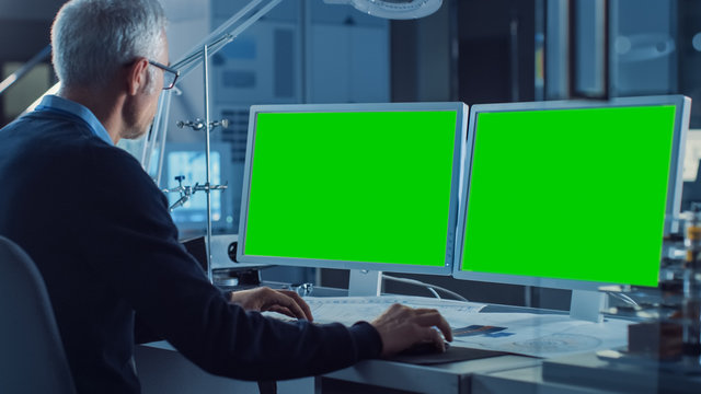 Professional Engineer Works On Computer With Two Green Mock-up Screen Displays. In The Background Industrial Electronics Design Agency. Over The Shoulder Shot