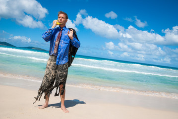 Castaway businessman standing in tattered suit using a banana as a mobile phone to have an imaginary conversation on a tropical beach