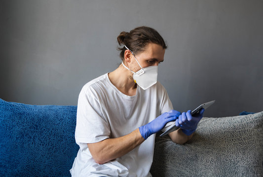 Man in white shirt, medical mask and rubber gloves sits at home and works with tablet on a sofa during quarantine. Man, designer, artist, architect, businessman at remote work in a pandemic covid