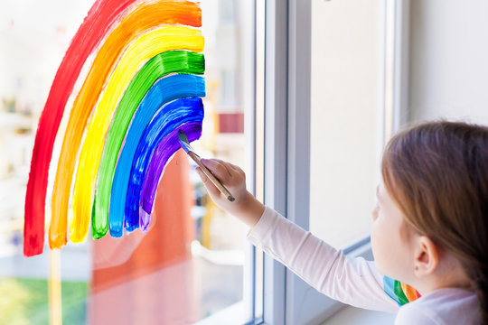 Kids at home. A child girl paints a rainbow on a window during the quarantine for the coronavirus pandemic. Social flash mob in support of society. Let's all be well. Stay at home
