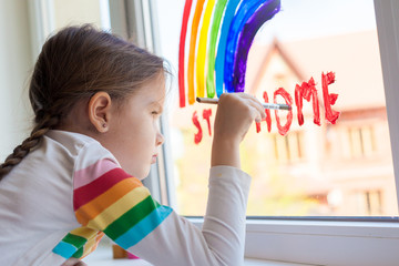 Kids at home. A child girl paints a rainbow on a window during the quarantine for the coronavirus pandemic. Social flash mob in support of society. Let's all be well. Stay at home