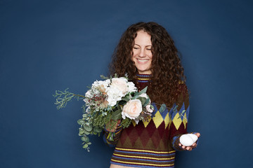 Studio image of curly haired female with perfect smile holding bouquet of plastic flowers and eggs, symbols of spring and Easter,  due to virus can not get fresh and natural, isolated on blue wall.