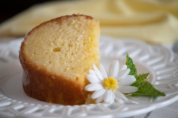 A slice of moist Lemon Ricotta Bundt Cake on a white decorative plate with a daisy and mint leaf garnish.
