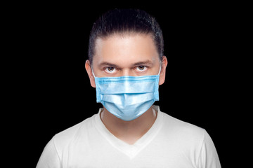 Male portrait with a serious facial expression of a medical worker in a white coat and blue medical protective mask and sterile gloves, isolated on a black background.