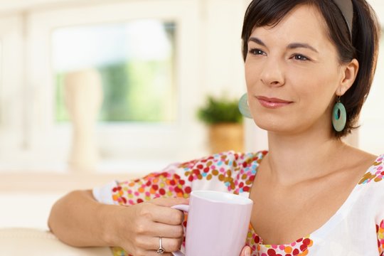 Portrait of young woman at home holding morning coffee cup, looking away, smiling.