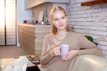 Beautiful young nordic type woman sitting on sofa at home, drinking tea, daydreaming.