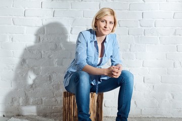 Casual young blonde woman sitting on stool front of white brick wall, smiling, looking at camera.