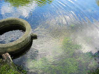 Fototapeta premium Concrete springlet with wooden path in small river surrounded by foliage. Well at countryside in summer time, reflection of blue sky and white clouds on the water
