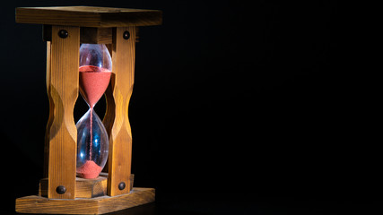 an old wooden hourglass with pink flowing sand in the left part of the frame. the lower part is...