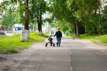 Fototapeta premium A small fair-haired boy leaves the sidewalk from the viewer. He leads his tricycle blue bicycle by the handle next to him. Lonely child, lost, fear, horror, hopelessness