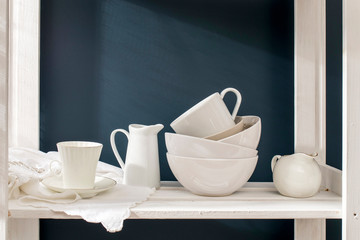 Fototapeta na wymiar A a set of white dishes: porcelain milk jug, coffee cup, bowls on a white rack on a dark blue background