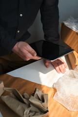 A man is packing cardboard boxes. Delivery service for online orders. Holding tablet in hand. The guy working in office, the cargo business. Warehouse with products, preparation for express shipping