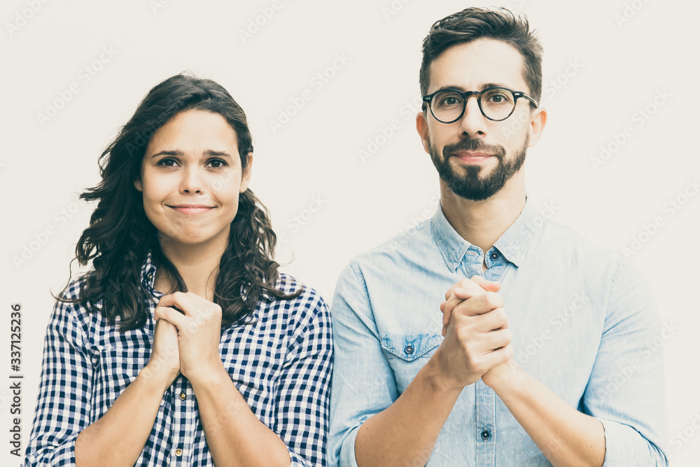 Wall mural Excited couple hoping for luck, making prayer gesture. Young woman in casual and man in glasses standing isolated over white background. Wish concept