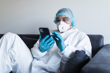 Young man in biohazard clothing using phone and staying at home at self isolation