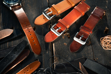 Men's luxury watch and handmade brown tone watch straps with steel buckles laying on the wooden rustic surface next to leather threads. Different leather colour tones.