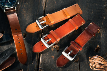 Leather brown watch straps with steel buckles laying on the dark wooden surface near to the leather thread. 