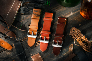 Handmade brown leather watch straps with steel buckle on rustic wooden surface next to leather cuttings and thread. Different leather colour tones. - Powered by Adobe