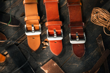 Close up of different colour handmade leather straps with steel buckles near to the on the leather thread on the dark wooden table. 