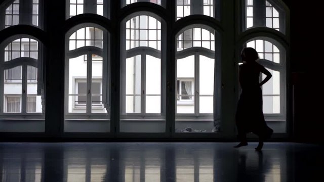 The silhouette of a girl in a long skirt , she is in a dark large room with large Windows and dances beautifully. He makes movements with his hands , whirls and skips. Against the light.