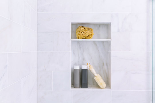 Bathroom shower interior with gray marble tile and shelves with shampoo and conditioner bottles, brush, and sponge 