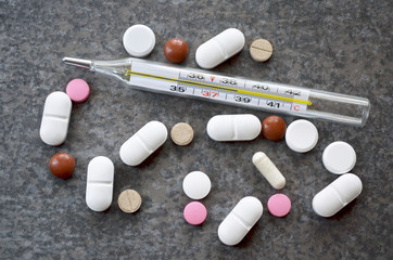 Colorful pills near to a medical mercury-in-glass thermometer indicating a critical temperature of 38.6 degrees Celsius on a dark gray table surface. Close-up, top view