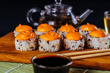 Delicious rolls on wooden board with chopsticks and green tea in teapot on decorative bamboo napkin. Close up of tasty sushi rolls on cutting board on black background.