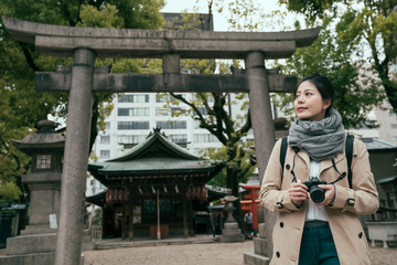 Asian woman tourist taking photos in tenmangu temple. young girl tourist traveling along osaka japan active lifestyle concept. beautiful lady holding camera with torii in background outdoors.