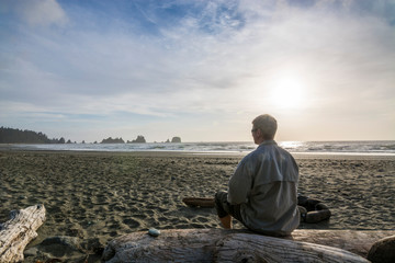 Adventurous man sitting on drift wood by a rugged Pacific Northwest beach watching the sunset.