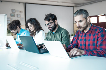 Line of focused coworkers sitting at meeting table with laptops. Business colleagues in casual working together in contemporary office space. Teamwork concept