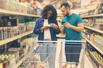 Serious young couple standing in aisle with alcohol drinks. African American people making purchases according to shopping list. Shopping concept
