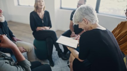 Group of corporate professionals laughing during team building training session in the office. Coach using a digital tablet with the multi-ethnic business team sitting in a circle.
