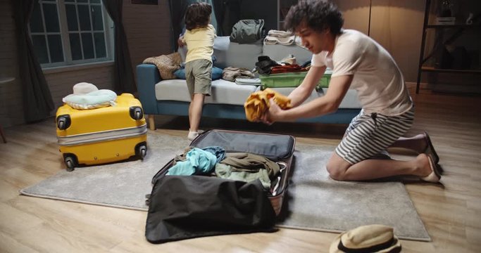 Funny asian young dad and preschooler son with curly hair are rapidly packing clothes in suitcase at home, preparing for their vacation. Family before road trip 4k footage