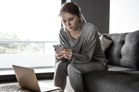 Concentrated sick young Caucasian woman in grey homewear sitting on grey sofa in living room, texting on phone. Laptop on table. Illness, work concept