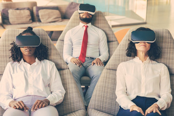 Focused business colleagues in VR headsets. High angle view of multiethnic businesswomen and businessman sitting in chairs and using virtual reality headsets. Technology concept
