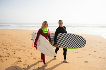 Happy surfers going away from beach and talking. Smiling man and woman holding surfboards and...