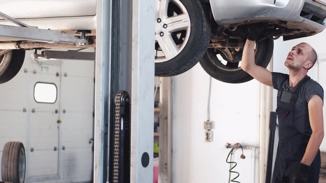 Worker wears uniform and gloves. Mechanic fixes car. Man works under machine.