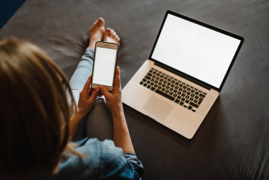 Girl checking social apps on white blank screen smartphone in home. Woman working and using laptop computer in the bedroom. Freelancer. Writing, typing. Communication and technology concept. Top view.