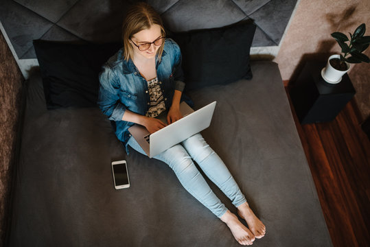 Beautiful young woman working, using laptop computer in bedroom. Freelancer. Writing, typing. Girl checking social apps. Communication and technology concept. Black blank screen smartphone. Top view.