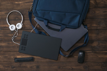 Graphic tablet, laptop and headphones sticking out of the bag on a wooden background.