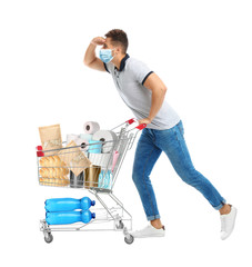 Young man in medical mask and shopping cart with purchases on white background