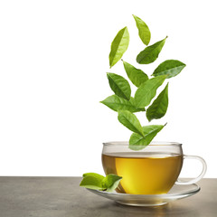 Green leaves falling into cup of tea on table against white background