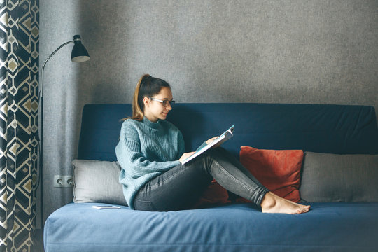 A girl reads an entertainment or educational magazine while sitting on a sofa in a cozy home environment.
