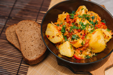 Stewed potatoes with cabbage in a dark plate on a brown board, on a light wooden table. Rustic cuisine.