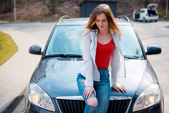 Young Stylish Woman In Red Tshirt With White Shirt And Fashionable Blue Jeans Sitting On A Hood Of New Expensive Car Outdoors. Woman Enjoying A Sunny Spring Day