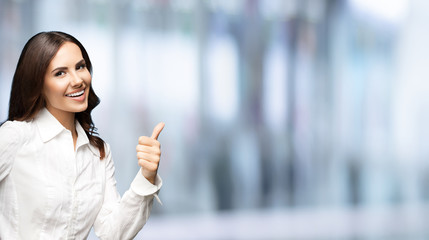 Fototapeta premium Portrait of happy smiling young excited businesswoman, showing thumb up hand sign gesture. Success in business concept studio shot. Standing against blurred office background.