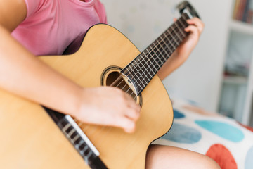 Cute tween girl in pink t-shirt play the guitar sit on bed in bright room at home