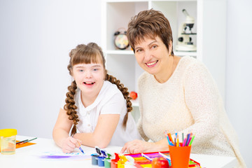 The girl and mother sitting at the table draw a picture at home. A girl with down syndrome at home...
